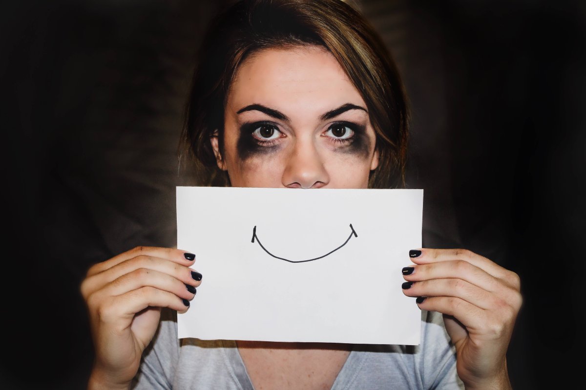 Photo of a lady who's makeup has been smeared from crying but is holding up a smiley face drawing over her mouth. 